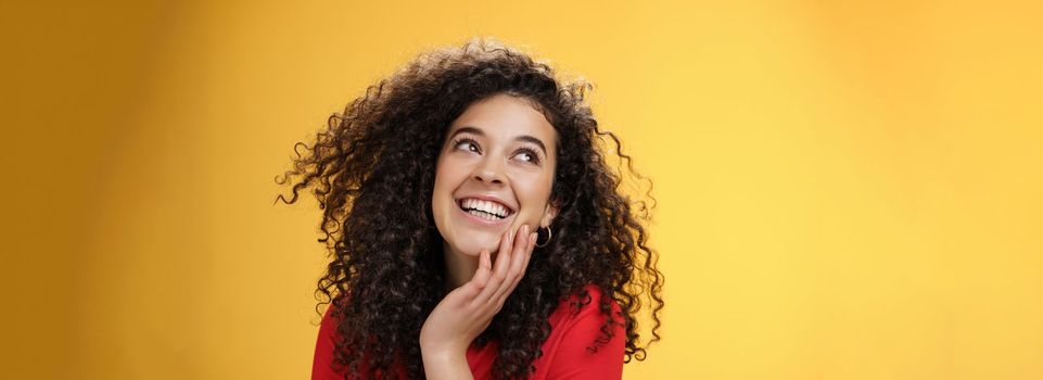 Happy positive pretty young woman with curly hair in red blouse laughing silly and carefree as gazing pleased at upper left corner, touching face, satisfied and delighted over yellow wall.