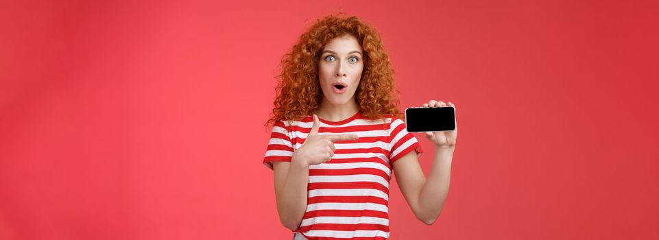 Lifestyle. Amused fascinated cute redhead curly girl adore playing games hold smartphone horizontal show thumb up open mouth gasping amazed happy cheerfully present own score favorite app red background.