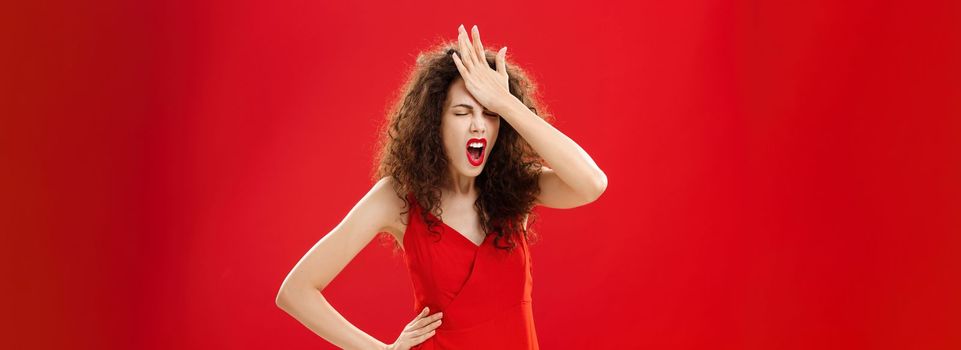 Woman being pissed with dumb people. Irritated forgetful adult caucasian female with curly hair in red elegant dress punching forehead with palm closing eyes and swearing recalling important thing.