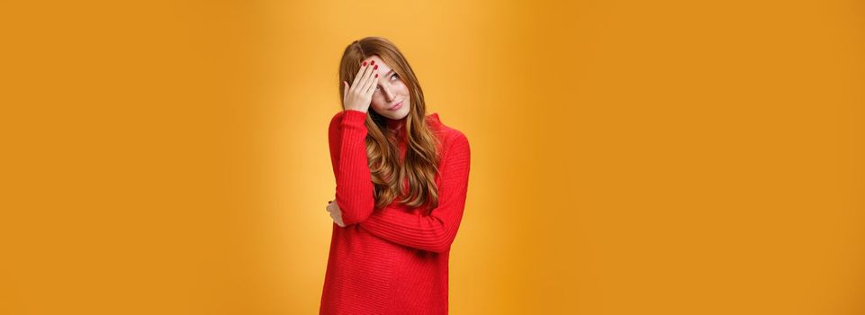 Annoyed and pissed embarrassed woman hiding behind hand making facepalm gesture looking away with irritated humiliated gesture, standing fed up and intense against orange background.