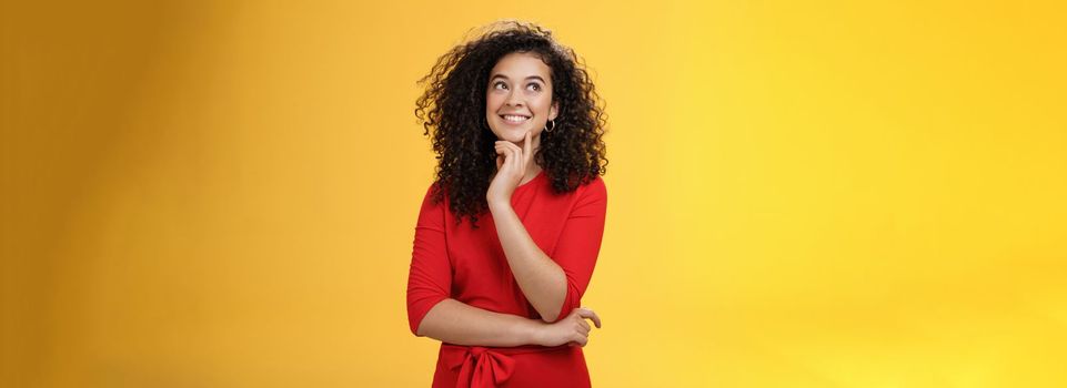 Girl having interesting idea, smiling as feeling confident plan might work. Charming happy woman with curly hair in red dress gazing at upper left corner thoughtful, thinking over yellow wall.