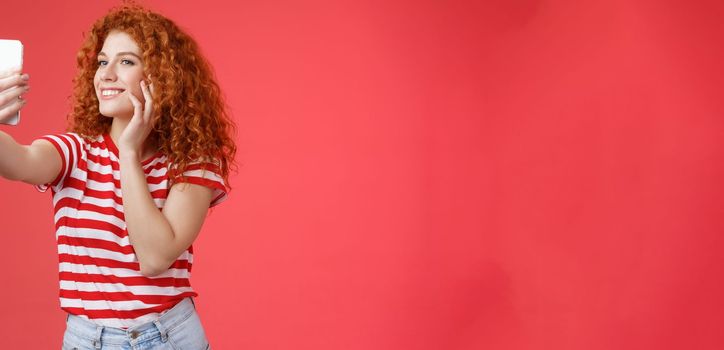 Lifestyle. Popular cheerful good-looking stylish female blogger redhead curly hairstyle feel pretty self-acceptance taking selfie raised arm holding smartphone posing silly cute phone camera red background.