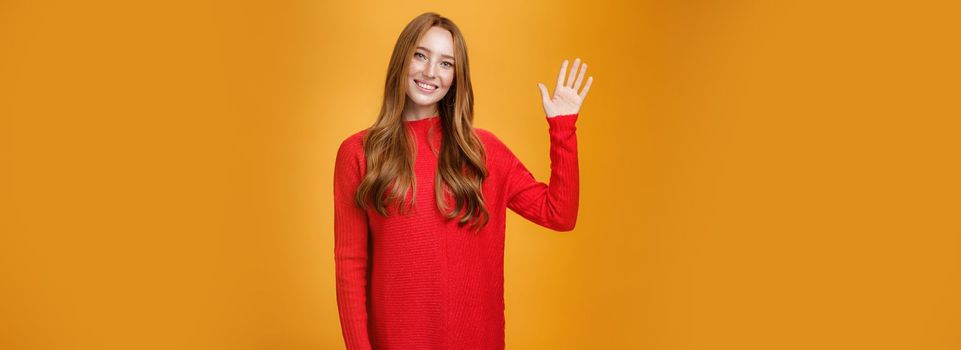 Friendly and optimistic good-looking ginger girl in red sweater raising palm waving at camera, saying hello or hi joyfully smiling cute, greeting new members posing over orange background.