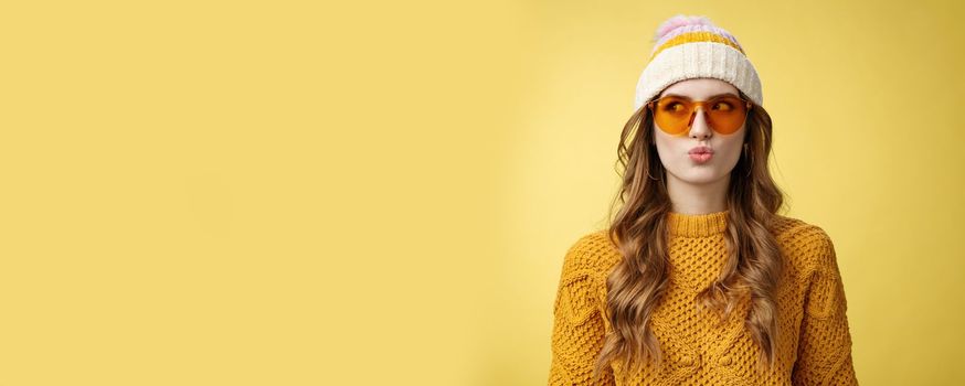 Stylish young glamour thoughtful girl make desicion folding lips silly thinking choosing product look away left wearing sunglasses hat warm sweater enjoying vacation ski resort, yellow background.