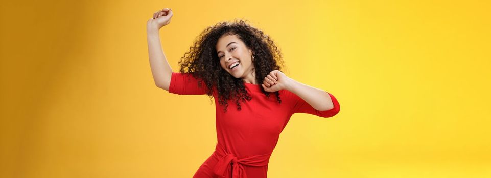 Carefree girl feeling uplifted and joyful dancing in red dress raising hands up happily tilting head and smiling broadly at camera as enjoying vacation, celebrating holidays over yellow background.