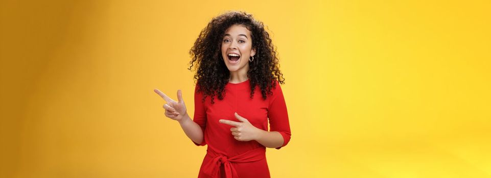 Thrilled and happy joyful young charismatic woman with curly hair in red dress laughing out loud and smiling amazed, delighted pointing at upper right corner impressed over yellow background.