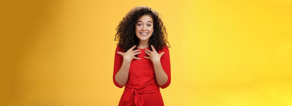 Lifestyle. Pleased and thankful tender curly-haired kind girl in red dress pressing hands to breast being surprised with unexpected tender heartwarming gift thanking smiling delighted over yellow background