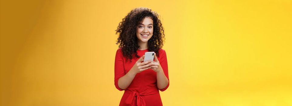 Girl messaging friend to tell all hot rumors holding mobile phone in hands smiling broadly and excited at camera as using smartphone browsing in net or edditing in app over yellow background. Technology concept