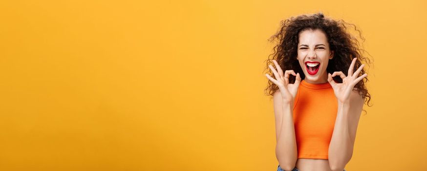 Charismatic happy and joyful attractive young female. with curly hairstyle showing okay gestures with both hands in approval or confirmation smiling and laughing happily over orange wall.