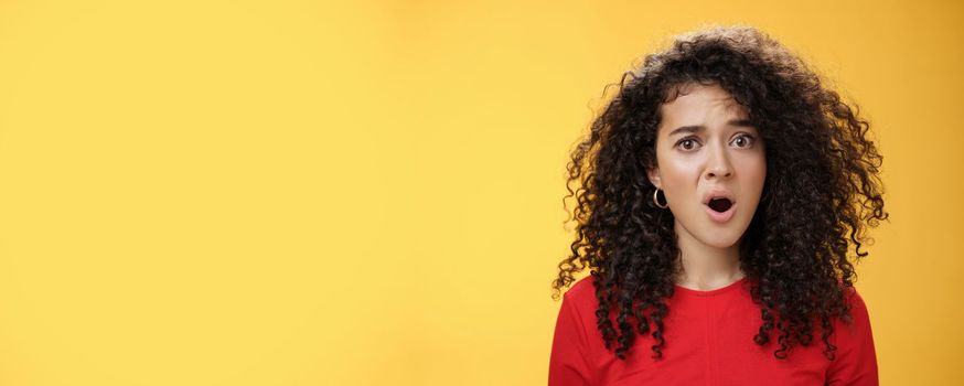 Lifestyle. Confused and frustrated young questioned woman with curly hair open mouth and raising eyebrow in surprise being displeased with unfair situation standing clueless and upset over yellow background.