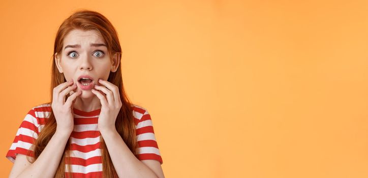 Scared shocked redhead woman worry, stare camera speechless gasping concerned, touch lips drop jaw astonished, hear terrifying scary news, stand orange background stupor, very upset.