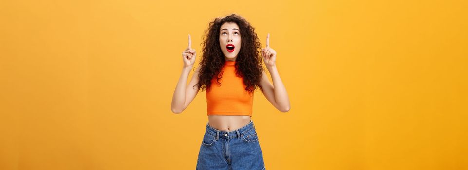 Portrait of amazed emotive and charismatic young curly-haired woman with red lipstic looking and pointing up mesmerized and astonished standing under impression over orange background. Copy space