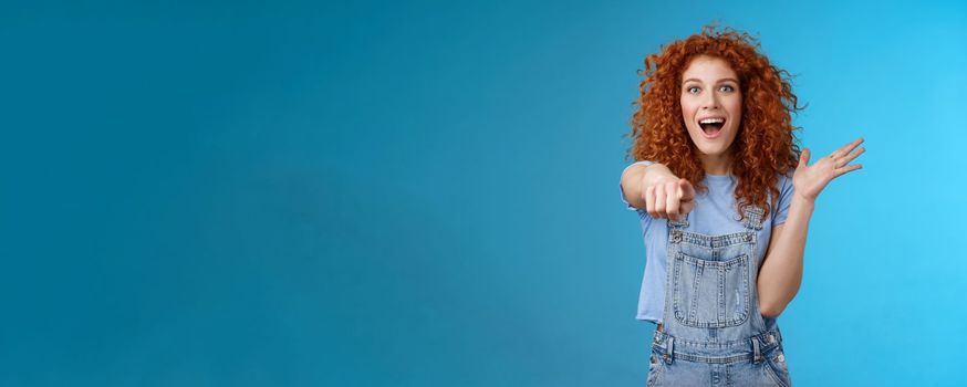 Excited impressed surprised cute redhead european woman curly astonished happily pointing index finger camera gesturing lively thrilled see awesome thing reacting amazed standing blue background.
