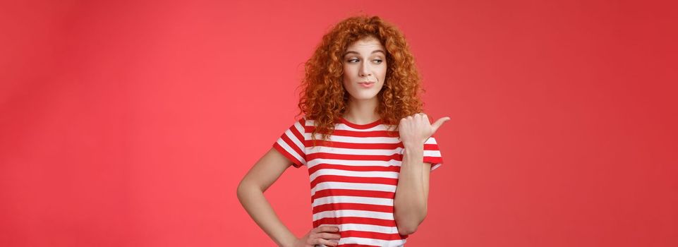 Look that stuff. Redhead curious sassy good-looking curly-haired girl summer striped t-shirt smirking gazing pointing left indicating thumb intriguing product standing red background.