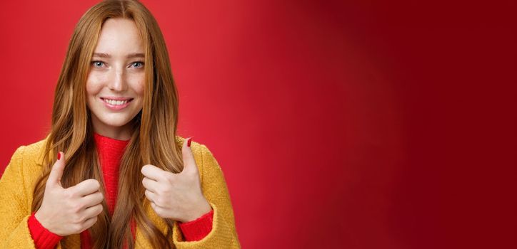 Close-up shot of satisfied happy young 20s woman in yellow coat showing thumbs up gesture in like and approval giving positive feedback enjoying nice event standing over red background, smiling cute.