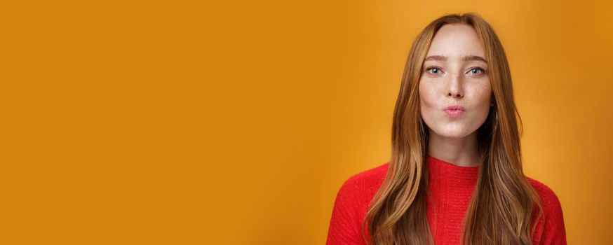 Close-up shot of flirty good-looking sensual redhead female in red knitted clothes folding lips in kiss and gazing at camera as giving mwah feleing romantic and pretty against orange background. Body language, fashion and people concept