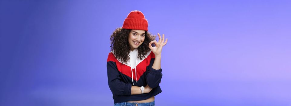 Portrait of pleased good-looking assertive woman with confident look and curly hair wearing warm winter hat showing okay gesture and smiling assuring, liking and approving awesome outfit of friend.
