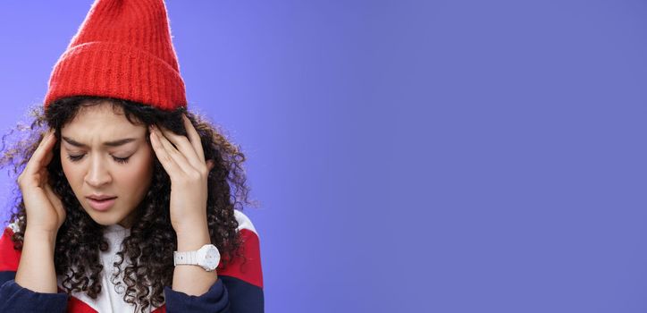 Close-up shot of sad and sick cute stylish woman in red warm beanie close eyes rubbing temples feeling tired and suffering headache as catching cold, standing with terrible migraine over blue wall.