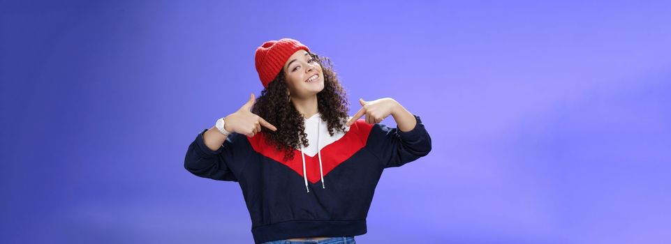 Lifestyle. Proud and self-satisfied happy young energized stylish female model in warm beanie brag about own achievements as pointing at herself raising head and smiling produly posing delighted over blue wall.