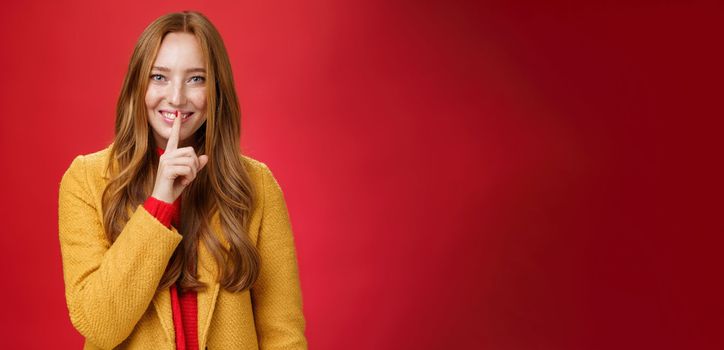 Shh keep it secret. Flirty and gentle attractive ginger girl with freckles teeling mysterious things showing shush gesture with index finger on mouth, smiling having surprise over red background.