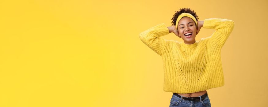 Carefree joyful lucky african-american female student in sweater headband lay back hands behind head relaxed chilling pose having day-off weekend smiling broadly laughing, yellow background.