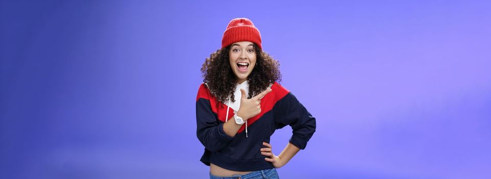 Lifestyle. Portrait of enthusiastic and sociable european woman in warm hat and sweatshirt smiling delighted with amused grin as pointing at upper left corner impressed and astonished with awesome promotion.