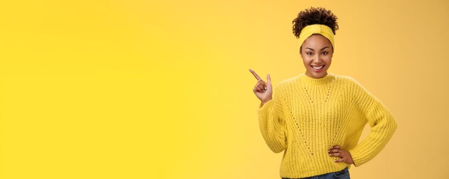 Charming sassy stylish african-american girl. in sweater pointing upper left corner standing confident cheeky energized smile yellow background promoting merch showing best choice look camera amused.