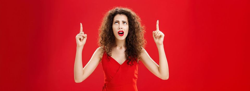 Troubled woman cannot understand what happening. Portrait of clueless silly european female with curly hair in red dress looking and pointing up perplexed and questioned posing over studio background. Copy space