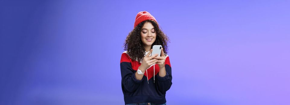 Woman searching in internet funny memes, holding mobile phone in hands smiling joyfully and cute at smartphone screen, typing message or browsing internet over blue background, wearing warm winter hat.