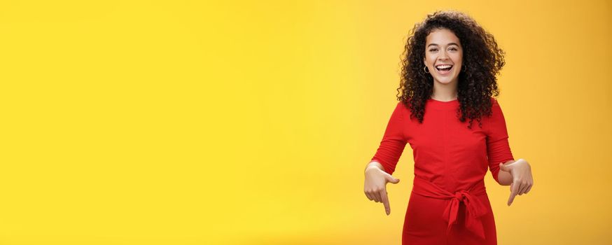 Waist-up shot of assertive charming and happy young woman with curly hairstyle laughing joyfully, smiling and pointing down as showing cool place to check out over yellow background. Advertisement concept