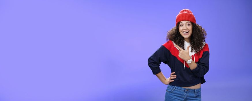 Lifestyle. Stylish and sociable friendly-looking attractive woman with curly hair in warm beanie pointing at upper left corner laughing from joy and amazement having fun posing happily over blue background.
