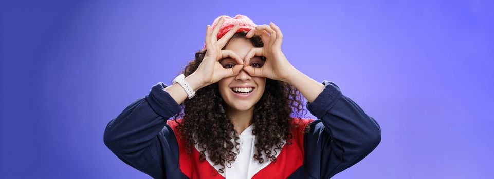 Girl see us as imitating peek with binocular making circles over eyes and looking through it joyful and excited, having fun fooling around posing happy and cute over blue background in outdoor outfit.