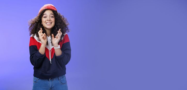 Optimistic and hopeful girl having faith in desire come true, standing with crossed fingers for good luck having wish anticipating miracle, smiling broadly at camera as wearing warm stylish red beanie.