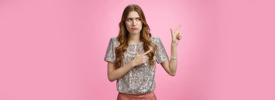 Displeased arrogant snobbish young rich glamour girl looking displeased cringe pointing left sideways, dislike place standing awkward insecure, expressing aversion reluctance, pink background.