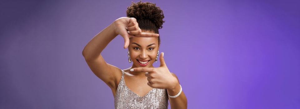 Creative good-looking african woman in silver glittering dress searching inspiration make finger frame look camera pleased smiling satisfied found perfect photo angle, standing blue background.