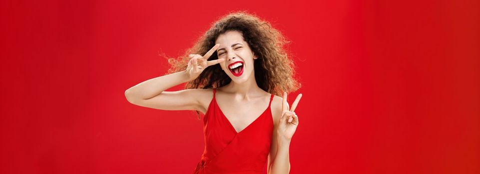 Outgoing friendly and carefree young rebellious woman with curly hairstyle in red stylish dress showing peace signs as if dancing disco enjoying awesome party having fun against studio background. Copy space