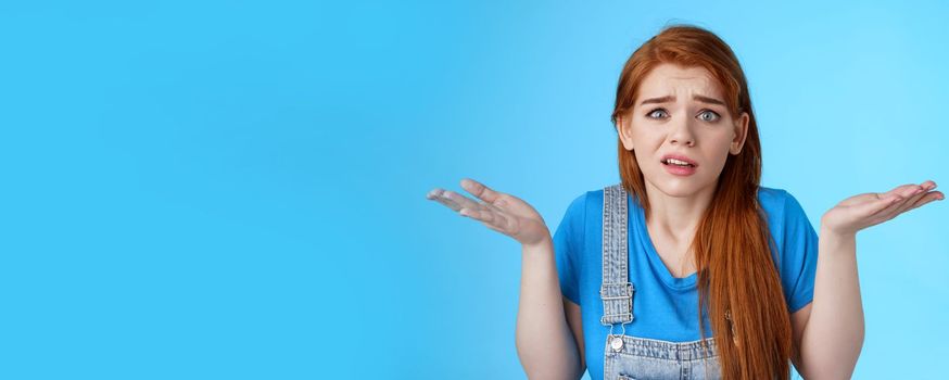 Perplexed uneasy stupified worried redhead woman shrugging, have no idea, frowning nervously, cannot understand what happened, stand clueless unsure, pose blue background. Copy space
