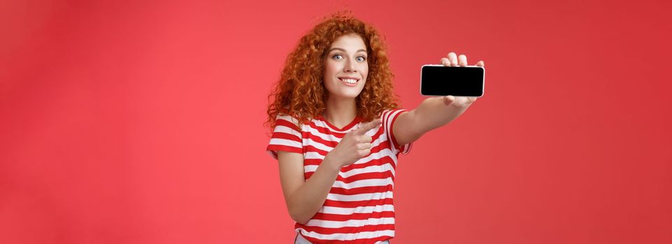 Excited happy good-looking redhead curly female show horizontal smartphone screen pointing display gadget smiling pleased proud beat friends score game standing red background. Technology concept