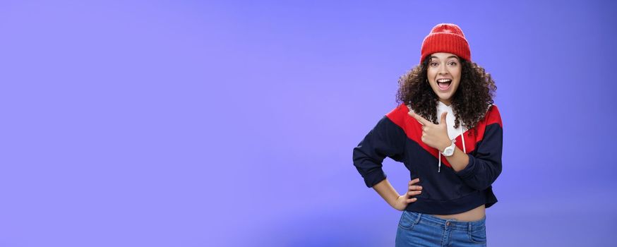 Lifestyle. Portrait of enthusiastic and sociable european woman in warm hat and sweatshirt smiling delighted with amused grin as pointing at upper left corner impressed and astonished with awesome promotion.