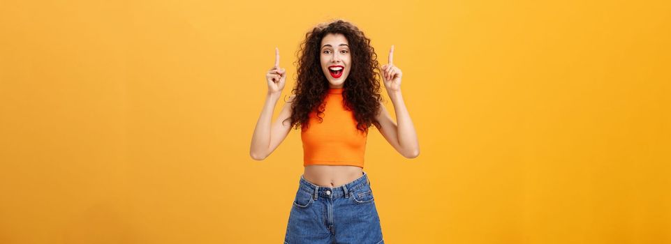 Woman surprising boyfriend with cool sign hanging upstairs pointing up, smiling singing happy b-day standing carefree and joyful over orange background in stylish cropped top and summer denim shorts. Copy space