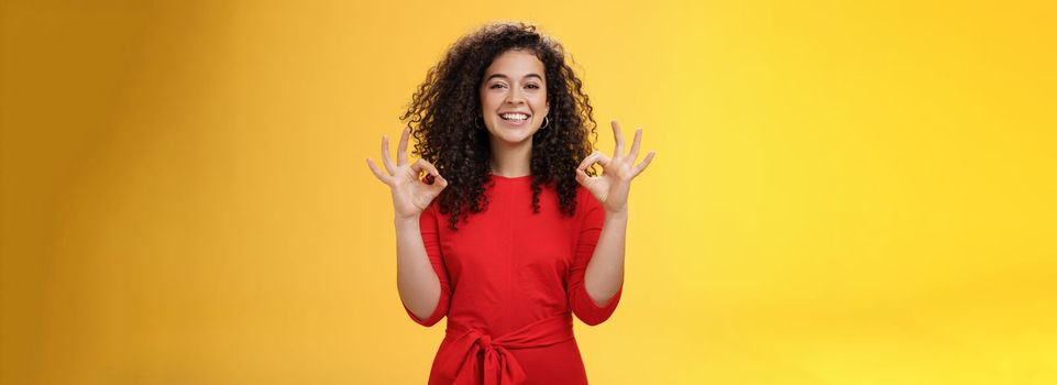Girl pleased everything goes on plan and okay. Happy joyful charismatic woman with curly hair in red dress showing ok gesture in approval and like, satisfied with good work over yellow wall.