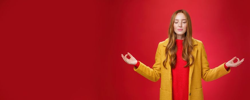 Girl keep calm releasing stress with meditation, posing in yellow coat, close eyes and looking relieved as extending hands sideways with mudra gesture, doing yoga against red background in lotus pose.