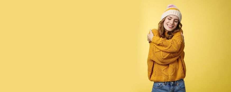Tenderness, happiness wellbeing concept. Charming feminine cute stylish girl liking new warm sweater hugging embracing herself close eyes dreamy smiling feeling romantic coziness yellow background.