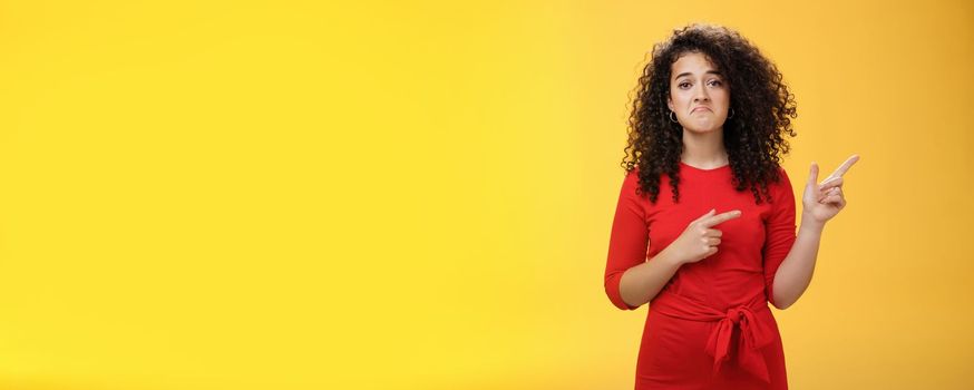 Waist-up shot of upset displeased girl with curly hair being down in dumps pointing at upper right corner with disappointed gloomy smile, feeling regret, sadness standing like loser over yellow wall.