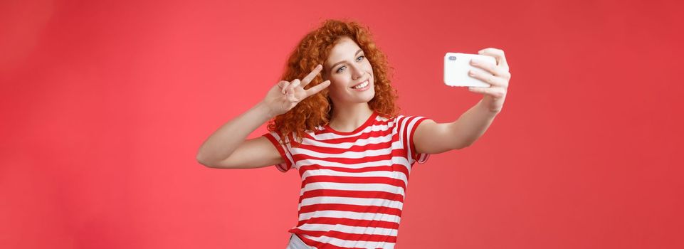 Stylish carefree good-looking redhead curly woman enjoy summer holidays show victory peace gesture eye tilt head cute record video hold smartphone taking selfie red background. Copy space