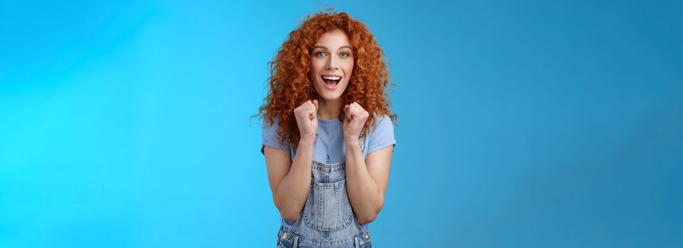 Cheerful optimistic lucky redhead curly-haired attractive woman cheering clench fists joyfully smiling broadly cheering watching game supportive encourage keep up motivated blue background.