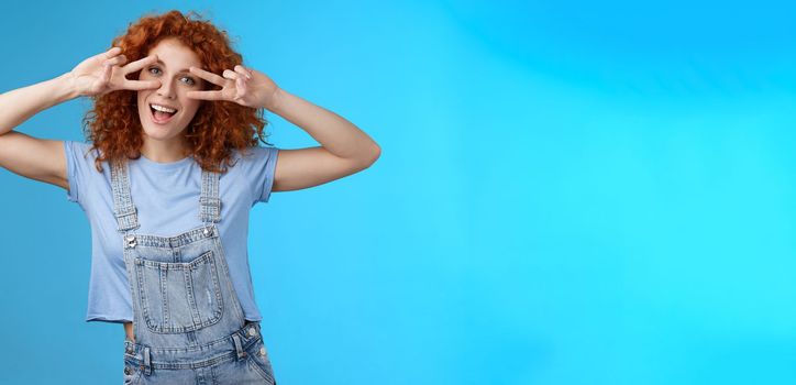Lifestyle. Cheerful cute redhead ginger girl curly haircut show positivity peace victory signs eyes smiling broadly have fun playful silly childish mood wear denim summer overalls blue background.