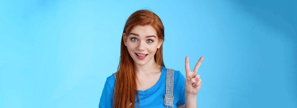 Cheerful attractive redhead caucasian girl smiling joyfully talking, order two peaces, show second number, peace victory sign, stand carefree, upbeat emotions, stand blue background.