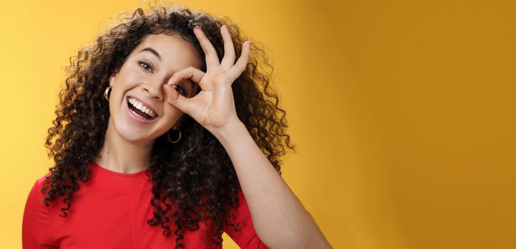 Hey I see you. Charismatic funny and playful cute tender girlfriend with curly hair showing okay or zero gesture over eye peeking at camera and smiling broadly, tilting head right over yellow wall.