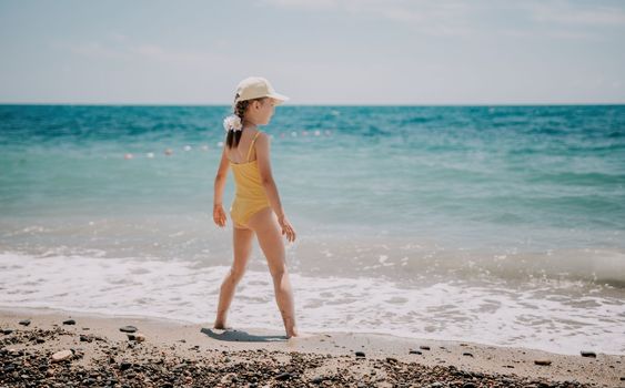 Cute little girl running along the seashore against a clear blue sea and rejoices in the rays of the summer sun. Beautiful girl in yellow swimsuit running and having fun on tropical beach